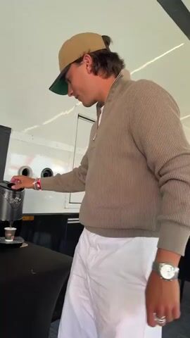 A man wearing a tan baseball cap backward, a tan quarter-zip sweater, and white pants is standing in front of a black table and a coffee machine. He is using his right hand to operate the coffee machine, which is dispensing a small cup of coffee. To the left of the coffee machine are several stacked paper cups. The man is also wearing a watch on his left wrist and several rings on his fingers. The background appears to be inside a white trailer or RV.