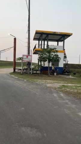 The video begins with a view of a gatehouse or guard post with a yellow roof, white walls, and blue trim. A red and white sign is visible on a pole in front of it. A road leads away from the gatehouse, with a large, tilting metal structure, possibly a gate arm, extending over the road. There are power lines and poles in the background, along with a grassy embankment and trees. The sky is light and overcast. The camera pans to the left, showing more of the tilting gate arm and the grassy area. The video continues to pan, revealing more of the environment, including a concrete barrier and pipes running along the embankment. The camera eventually stops panning, showing a wide view of the road and the surrounding landscape. The scene appears to be an industrial or utility area. There are no people clearly visible in the video, though there may be a figure in the gatehouse. The lighting suggests it is early morning or late evening.