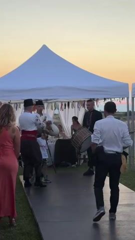 A group of people are gathered at an outdoor event under a white tent. A man in traditional clothing plays a bagpipe-like instrument while another man plays a drum. A small boy in a white t-shirt and green shorts stands near the man with the bagpipes, seemingly interacting with him. Other people are visible in the background, including a woman in a long pink dress and a man in a white shirt and black pants facing away from the camera. The sky in the background is a warm orange and yellow, indicating either sunrise or sunset. The event is set up on a grassy area, with a pathway leading to the tent.
