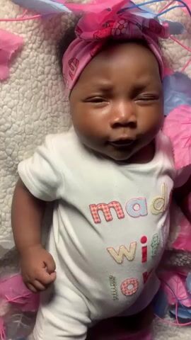 A close-up video shows a baby lying on a white, textured surface, possibly a blanket or playmat. The baby, with dark skin and a full head of dark hair, is wearing a white short-sleeved onesie with the embroidered text "mad with love" in colorful, checkered letters. A pink headband with a bow and a pattern resembling strawberries adorns the baby's head.

The baby's eyes are mostly open, occasionally blinking or closing briefly. The expression is calm and curious, with soft features. Around the baby are scattered pink and light blue leaf-shaped decorations, some connected by thin wires, creating a decorative frame. In the latter part of the video, the baby's left hand reaches out and gently touches one of the pink leaf decorations. The lighting appears to be bright and even, suggesting an indoor setting during the daytime.