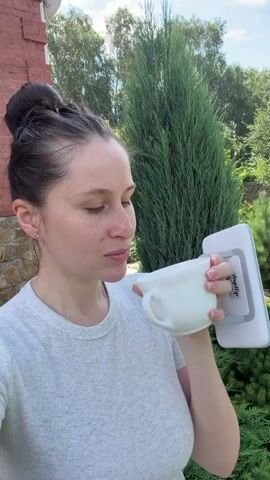A woman with her hair pulled back in a bun is drinking from a white mug. She is wearing a grey t-shirt. She is standing outdoors in front of a building and some trees. A white object with a handle is attached to the back of the mug. She takes a sip and then looks at the camera and smiles.