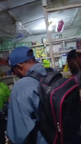 The video shows a crowded indoor space, possibly a small store. There are several men present. One is wearing a blue cap and a gray shirt with a black backpack that has red accents. Another man has a haircut with visible waves. A third man smiles at the camera, wearing a light green shirt and a cap. The environment appears cluttered with shelves stocked with various items. The lighting is somewhat dim. The video captures a casual interaction within the space.