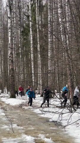 The video depicts a group of people running through a snowy, wooded area. The runners are wearing various athletic clothes and racing numbers. They are navigating a muddy patch of snow and ice. The forest is filled with birch trees. Some runners are helping each other over fallen branches. The sky is overcast, and the lighting is dim.