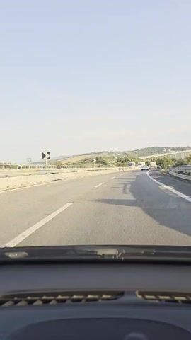 The video is a point-of-view shot from inside a vehicle driving on a highway. The road is paved and has lane markings. The sky is clear and blue, and the sun is shining, casting shadows on the road. On the left side of the road, there is a concrete barrier and then a sloped embankment with some vegetation. In the distance, there are hills with trees and some buildings. On the right side of the road, there is another concrete barrier and a bridge structure. There are other vehicles on the highway, including a few trucks and cars. The video appears to be shot during the daytime.