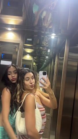 The video shows two young women taking a selfie in what appears to be an elevator. The woman on the right is holding a smartphone and taking the picture, while the woman on the left is leaning in and making a pouty face. Both women are wearing swimwear, suggesting they may be on vacation. The elevator has mirrored walls and wooden accents, and the lighting is bright. In the reflection of the mirror, another person is visible, possibly taking a photo of the two women. The scene is casual and playful.