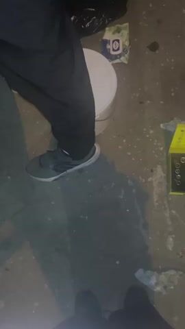 The video shows a ground-level view of an outdoor scene. A person wearing dark pants and gray athletic shoes stands with their legs partially visible, casting a shadow. Next to them is a white bucket. To the right of the bucket is an open yellow toolbox containing various metal sockets and tools. There are also some discarded paper or cloth items on the ground, as well as some dark splatters, possibly oil or dirt. The ground appears to be concrete or asphalt. The lighting suggests it is nighttime or a dimly lit area. The perspective is looking down from a slight height.