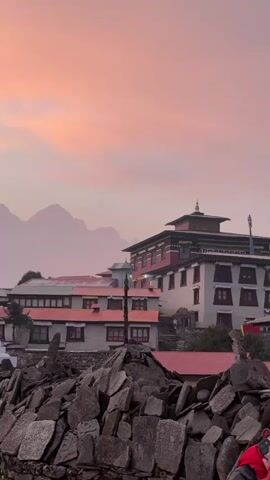 The video captures a serene and majestic Himalayan landscape, likely at either sunrise or sunset, characterized by a sky painted in soft pink and orange hues. The scene unfolds in what appears to be a traditional village, with several buildings featuring red roofs and white or light-colored walls, suggestive of monastic or residential structures. In the foreground, a low stone wall, possibly composed of mani stones (prayer stones), frames the bottom of the shot.

As the camera slowly pans from right to left, a prominent white stupa with a golden spire comes into view, standing tall as a focal point in the mid-ground. Behind the buildings and the stupa, towering, rugged mountains are silhouetted against the vibrant sky, their peaks partially obscured by a gentle atmospheric haze, emphasizing their grandeur. Dark green trees are scattered across the landscape, adding depth to the scene. The overall impression is one of peacefulness and natural beauty, characteristic of a high-altitude Buddhist settlement like Tengboche, Nepal. No people or discernible text are visible in the video.