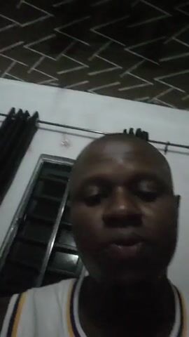 The video shows a close-up shot of a Black man, seen from the chest up, speaking directly to the camera. He has a shaved head and is wearing a white sports jersey with yellow and purple trim around the neck and armholes. The background includes a white wall, a black curtain, a window with visible metal bars, and a patterned ceiling with rows of zig-zag lines in shades of brown. The man appears to be indoors.