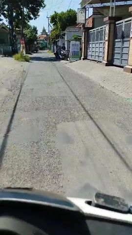 A first-person perspective video shows a person riding a motorcycle down a narrow paved street. On the right side of the street, there are houses with large metal gates and some trees. On the left side of the street, there are more houses and some vegetation. There are a few other motorcycles parked on the side of the road, and several people are standing or sitting around them. The sky is blue and the sun is shining, casting shadows on the road. The video is shot in daytime.