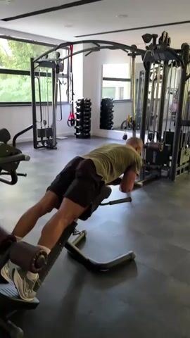 The video shows a man exercising in a gym. He is lying face down on a hamstring curl machine, with his legs extended. He is wearing a green t-shirt and black shorts. The gym appears to be well-equipped with various weight machines and equipment. Natural light is entering the room through large windows.