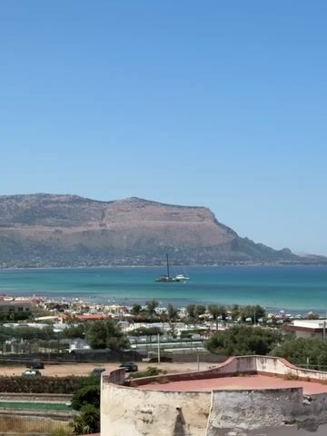 This is a landscape video capturing a serene coastal scene in Isola delle Femmine, Italy. The primary subjects are two sailboats anchored in the clear, turquoise waters of the Mediterranean Sea. One sailboat, a catamaran, is larger and more prominent, with a dark mast reaching towards the sky. A smaller sailboat is positioned behind it.

The scene is set against a backdrop of a rugged, mountainous coastline, with a small town nestled along the shore. The town features a beach dotted with umbrellas and beachgoers, suggesting a popular tourist destination. Lush green trees and vegetation are scattered throughout the foreground and mid-ground, adding to the natural beauty of the location. In the foreground, a partially visible rooftop with red tiles and white plaster hints at a building or dwelling from which the video is being filmed.

The activity taking place is passive observation of the seascape. The sailboats appear to be at rest, enjoying the calm waters. The presence of people on the beach indicates it is a warm, inviting day for recreation.

Notable details include the bright, clear blue sky, indicative of fair weather and likely a sunny day. The time of day appears to be midday, given the intensity of the sunlight. The vibrant blue and green hues of the sea are striking, contrasting with the earthy tones of the mountains. The overall mood is tranquil and picturesque, evoking a sense of vacation and relaxation. There is no visible text in the video.