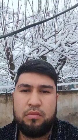 A man with short dark hair and a beard is shown in a close-up, facing the camera. Behind him are snow-covered trees and a partially visible wall. The man's expression is neutral.  It appears to be snowing.