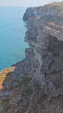 The video shows a rocky cliff overlooking a serene, turquoise sea under a clear blue sky. The camera pans across the scene, revealing the rugged texture of the cliff edge, which is covered in patches of orange lichen and sparse vegetation. The sea appears calm with faint ripples on the surface, and large, dark rocks are visible just below the waterline. In the distance, another cliff formation can be seen jutting out into the water. The overall impression is one of natural beauty and tranquility.