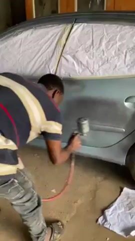 A man wearing a short-sleeved, horizontal striped shirt and pants is spray-painting the side of a blue car. The car's windows and roof are covered with white plastic sheeting, held in place with yellow tape. The man holds a spray gun attached to a red hose. The car is in what appears to be a garage or workshop with a concrete floor and a white cloth on the floor near the front tire. In the background, there's a wooden frame or door. The man is focusing on evenly coating the car's surface with paint.