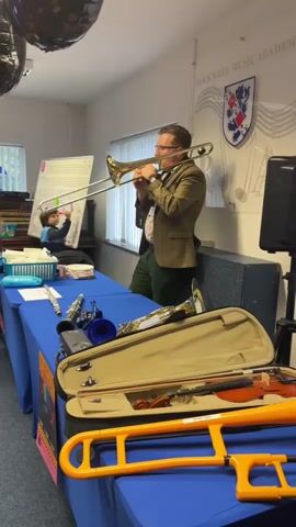 A man wearing glasses, a brown tweed jacket, and green pants is playing a trombone. He is standing behind a table covered with a blue tablecloth. On the table are various musical instruments, including a violin in a case, a flute, a blue trumpet, and a yellow plastic trombone. A young boy is sitting at the table, facing the man. In the background, there is a whiteboard with writing on it, and a logo that reads "ROCKWELL MUSIC ACADEMY". There are also black balloons hanging from the ceiling. The room appears to be a classroom or studio.