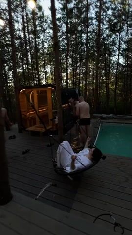 The video begins with a blurry view of a wooden deck. A person in a white robe is partially visible in the foreground, lying on a circular hammock-like object. To the left, a modern wooden sauna structure is visible, illuminated from within. Two shirtless men in dark shorts stand near the sauna. A rectangular swimming pool with green water is to the right of the deck. The scene appears to be outdoors, surrounded by trees, suggesting a forest or wooded area. The lighting indicates it is either dusk or dawn. The camera movement is shaky and fast-paced, creating a disorienting effect. Later shots show a person in a white robe sitting on the wooden deck, interacting with the camera.