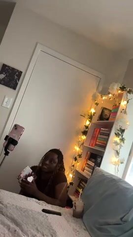 A medium shot depicts a person with long, light brown braids sitting on a bed and speaking to a smartphone held by a selfie stick. The person is holding a small, decorative object that appears to be wrapped in ribbon or lace, and holding it up toward the camera. Beside the person is a tall bookshelf with warm-toned lights strung across it, embellished with white flowers. A beverage with a straw sits on the bed next to the person.  Behind the person is a closed white door and a framed artwork on the wall above. The room is tidy.