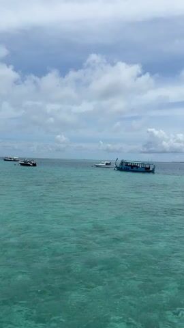 The video shows a view from a wooden pier over turquoise water. Several boats are visible in the distance, as well as an island with palm trees and buildings. The sky is cloudy. There are two people walking on the pier in the distance.