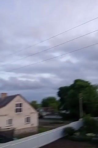 The video shows a view of a residential area under a dramatic, cloudy sky at dusk. The camera pans slowly to the right, revealing houses with various roof styles and colors. Power lines stretch across the top of the frame. There are trees and grassy areas between the houses. A single car is visible in the lower part of the frame. The lighting is dim, with a hint of orange on the horizon.