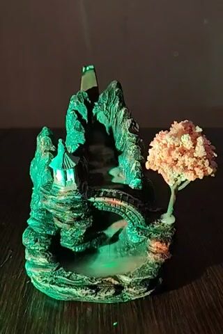 The video shows a close-up of an incense burner designed to resemble a miniature mountain landscape with a pagoda and a tree. Smoke is seen rising from the top of the mountain, cascading down in a waterfall-like effect. The scene is illuminated by changing colored lights, primarily green, blue, and purple, which cast an ethereal glow on the object and the surrounding surface. Small, scattered green lights are visible in the background, possibly reflecting from the colored illumination or part of the setup. The incense burner is placed on a dark wooden surface. The video appears to be a still shot with subtle lighting changes and no discernible movement of people or any other objects.