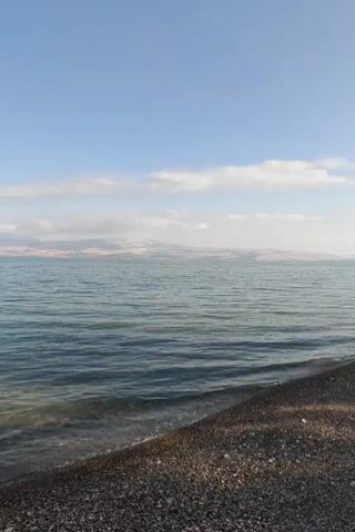 The video shows a body of water with a shoreline in the foreground. The water is a deep blue-green and appears calm with small ripples. The shoreline is composed of small, dark pebbles. In the distance, there are hills or mountains, and a hazy, pale blue sky with a few wispy clouds. The lighting suggests it is daytime, possibly morning or late afternoon, with the sun not directly visible but providing ample illumination. There are no people or distinct objects visible in the frame other than the natural landscape.