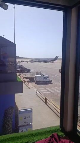 The video shows an airplane parked on a tarmac at an airport. The plane is white with a blue and gray stripe, and it says "Aerotucana" on the side. There are several people standing near the plane, presumably passengers or crew.  A white "Utaca" van is parked near the airplane. The view is seen through a window, where departure gate signage is visible. The weather is sunny and clear.