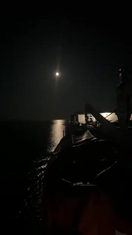 A drone shot from a dark, choppy ocean surface shows a large ship at night. The full moon is high in the sky, casting a bright reflection on the water. The ship's deck lights are on, illuminating a section of the vessel. The camera pans slightly to the left, revealing more of the ship's dark hull and the water churning behind it.