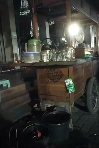 The video shows a street food cart at night. The cart is made of wood and has a wheel on the right side. There are several glasses and kettles on the counter of the cart. A light illuminates the cart from above. A person is sitting on a stool behind the cart, and appears to be serving a customer or preparing food. There are also buckets on the ground in front of the cart. The background shows a building and a street.