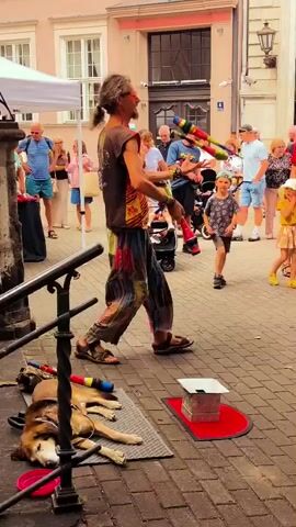 A long-haired, middle-aged man with glasses and a colorful patterned shirt and pants is juggling five clubs in an outdoor urban setting. He stands on a small platform in front of a red mat, with a dog sleeping on a mat to his left. A group of people are watching him, and some are walking by in the background. A baby is in a stroller near the juggler. The ground is paved with cobblestones, and there are buildings in the background. The overall impression is a street performance on a sunny day.