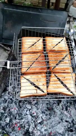 The video shows six triangular grilled cheese sandwiches on a metal grill basket over glowing coals. The sandwiches are golden brown and appear to be toasted on both sides. The grill basket is made of silver metal and has a handle on each side. The coals are red and orange, indicating that they are hot. The background of the video is blurry and appears to be an outdoor setting. The video is shot from a slightly elevated angle looking down at the sandwiches.