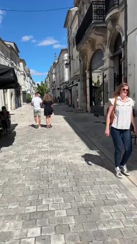 A wide cobblestone street is lined with buildings on both sides. The buildings are mostly two or three stories tall with arched windows and balconies. The street is shaded by the buildings, but the sky is blue with a few white clouds. Two people, a man and a woman, are walking away from the camera down the street. The man is wearing a white t-shirt and shorts, and the woman is wearing a black dress. There are a few other people walking in the distance. There are some tables and chairs set up outside a cafe on the left side of the street. There is a large potted plant with tropical leaves in the middle of the street. The video appears to be taken during the day.