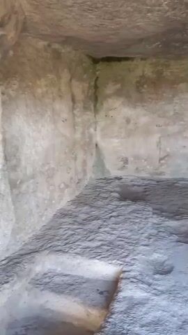 The video pans across the interior of a rough-hewn cave or rock shelter, showing its textured walls and floor made of dirt and stone. The camera moves to the opening of the shelter, revealing a view of a lush, green hillside under a bright blue sky with white clouds. A low, stone wall is visible on the hillside, and some greenery grows near the cave entrance.