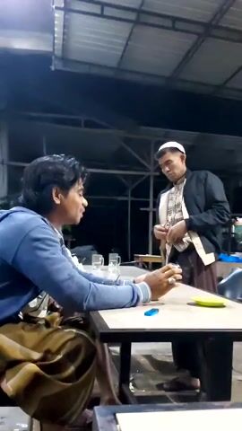 A low-angle shot captures two men at an outdoor café setting, under a metal-roofed structure. The man on the left, seated at a table, wears a blue long-sleeved shirt and a gold-toned sarong. He looks engaged with his mobile phone. On the table in front of him, there are clear glasses and a small blue object. The other man, wearing a white kufi cap, a patterned shirt, brown sarong, and a black jacket, stands opposite him, apparently adjusting his clothing. The background includes additional tables, chairs, and a food preparation or serving area with red and white elements. The setting suggests a casual evening atmosphere.