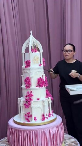 A medium shot shows two women about to cut a large multi-tiered white cake decorated with pink flowers, set against a pink curtain backdrop. The woman on the left, wearing a black shirt and glasses, holds a thin knife or sword and appears to be instructing or coordinating with the other woman. The woman on the right, also in a black shirt, holds a tray with plates. The cake is elaborate, featuring architectural details like columns and a birdcage-like structure on top, with gold lettering that says "Happy Birthday Stella". The cake sits on a pink-draped round table. The setting appears to be indoors with professional lighting.