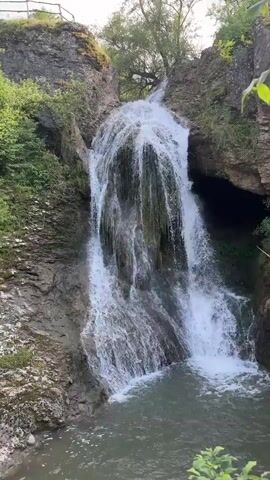 The video shows a waterfall cascading into a pool of water. The surrounding landscape includes rocky cliffs covered with vegetation and a cave entrance. A wooden walkway with a handrail is also visible.