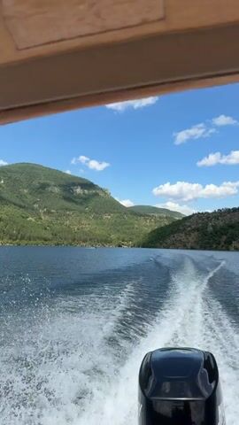 The video shows a boat moving across a body of water, leaving a wake behind it. The boat appears to be an outboard motorboat, as the motor is visible at the stern of the boat. The water is blue and calm, with some small waves. The surrounding landscape is mountainous and covered in green vegetation. The sky is blue with some white clouds. The video is shot from the perspective of someone sitting in the boat, looking backwards.