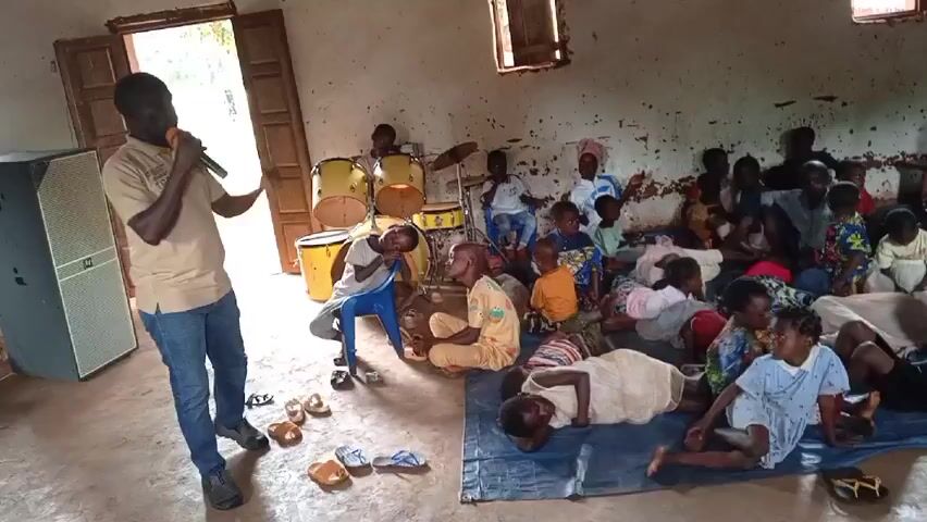 Inside a room, a man speaks into a microphone while standing in front of a group of people. The room appears to be old, with white walls and small windows. The floor is a reddish-brown color.

A large group of people, mostly children, are seated on the floor or on chairs, and a few children are lying on a blue tarp. There is a set of drums in the corner of the room. The children are wearing a variety of clothing, and some of the women in the back are wearing headscarves.

The man speaking is wearing a tan shirt and blue jeans. He is holding the microphone in his right hand and gesturing with his left. The people in the room appear to be listening to him. The scene seems to be taking place in some kind of community or religious setting.