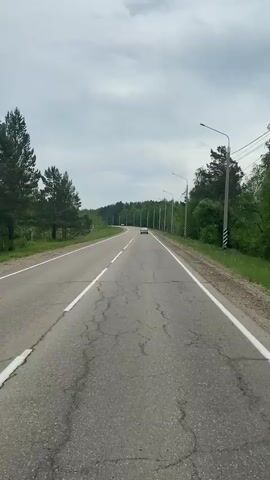The video shows a view from a moving vehicle on a paved road. The road has white lines marking the lanes and is cracked in several places. On either side of the road, there are trees and vegetation. There are also utility poles with lights and wires running along the right side of the road. A white car is visible in the distance, driving in the same direction. The sky is cloudy.