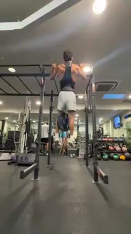 A man is performing weighted pull-ups in a gym. He is wearing a black tank top, grey shorts, and white sneakers. A weight plate is attached to his waist with a belt. He pulls himself up, chin over the bar, and lowers himself with control. The pull-up bar is set up within a black power rack. Other gym equipment is visible in the background, including kettlebells, cable machines, and a squat rack. Another person is visible working out further in the background. The gym has bright lighting from overhead fixtures.