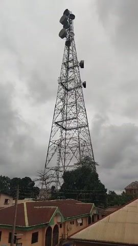 The video shows a telecommunications tower against a cloudy sky. The tower is a lattice structure made of metal. Several antennas are mounted at the top of the tower. Below the tower, the roofs of several buildings are visible, along with some trees. The camera pans upwards and slightly to the right, causing the telecommunications tower to fill more of the frame. The video ends with a blurred image of the sky and treetops.