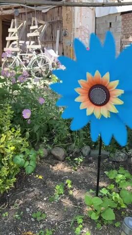 The video shows a garden scene with various plants and decorative items. In the foreground, a large blue pinwheel shaped like a flower with a sunflower center is prominently displayed. To the right of the pinwheel, tall lilies with pink and orange flowers are in bloom. Behind the lilies, a thin tree trunk with a twisted branch is visible. The background reveals a rustic setting with a wooden structure, some cinder block walls, and a bicycle parked behind a wooden fence. The ground is covered with dirt, small stones, and various green plants. The lighting suggests it is daytime with sunlight filtering through the leaves. The camera pans slowly to the right, capturing different parts of the garden.
