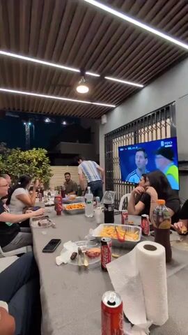 A group of people are gathered around a table outdoors at night, watching a soccer game on a large television screen. The table is covered with a grey tablecloth and set with food, drinks, and paper napkins. A man wearing an Argentine jersey is standing by the table, and other people are seated, eating, and talking. The atmosphere appears to be casual and celebratory.