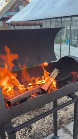 A charcoal grill burns with a healthy fire outdoors. Two pieces of firewood are laid across a bed of charcoal. The fire has created tall flames that reach up into the body of the grill. The grill is made of black painted metal and has a hinged hood. It is set on a black metal stand. The ground is covered with dry, patchy grass. Behind the grill is a residential setting with a green house, and a house with a wooden roof. The day is bright and sunny.