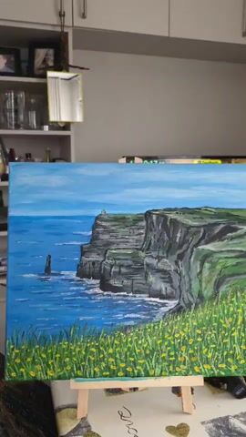 This is a video of a painting on an easel, likely a depiction of the Cliffs of Moher in Ireland. The painting features dramatic, green-topped cliffs plunging into a deep blue, choppy sea. A tall, slender sea stack rises from the water to the left of the cliffs. The foreground is filled with tall, green grass and small yellow flowers, suggesting a bright, possibly sunny day. The sky is a clear, pale blue. In the background, a small lighthouse or structure can be seen perched atop the cliffs. The overall impression is one of a rugged, natural landscape. The painting is displayed indoors, with shelves and cabinets visible behind it, suggesting it's in a home or studio. There is a digital display in the background showing "10:17:12," which could be a clock or a timer. The painting itself is the primary subject.