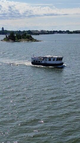 The video shows a ferry moving through the water near Helsinki, Finland. The ferry is white with a dark hull and has several decks with windows. It appears to be a passenger ferry, and there might be people visible on the decks, although they are not clearly discernible.

The scene is set on a body of water, likely a bay or channel, with a shoreline visible on the left side. The shoreline is lined with trees and some buildings, suggesting a populated area or town. A small, rocky island with some vegetation is also present in the water near the shore. The water itself is choppy and reflects the sunlight, creating a shimmering effect. The sky is partly cloudy, with a mix of blue sky and white clouds.

The ferry is in motion, leaving a wake behind it as it travels through the water. The activity taking place is transportation by ferry. The time of day appears to be daytime, given the bright sunlight and shadows. The weather seems fair, with sunshine and some clouds. The location cues (Helsinki, Finland) and the type of ferry suggest a scene common in coastal Finnish cities. There is no visible text in the video. The overall mood is peaceful and serene, typical of a day spent on the water.