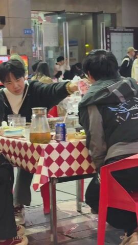 A group of people are seated around a table in an outdoor setting. The table is covered with a red and white checkered tablecloth. On the table, there is a pitcher of liquid, a can of soda, dishes with food, and various other items. One person is holding up a smartphone, displaying a picture of a person to another individual at the table. Other people are visible in the background, some seated at other tables and some walking. The surrounding environment appears to be a commercial area with storefronts and signage.