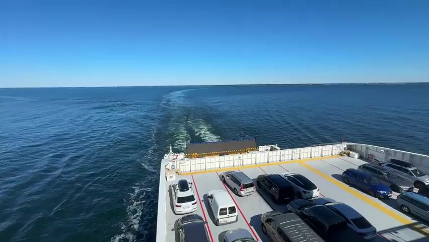The video shows the stern of a ferry with cars parked on its deck. The ferry is moving through calm blue water under a clear blue sky, leaving a wake behind it. The camera angle is high and looking down on the deck and the water. The cars are parked in rows on the open deck of the ferry. The water is a deep blue and appears calm, with the sun reflecting off its surface. In the distance, the horizon is visible, with a faint line of land or a coastline. The overall scene suggests a peaceful and clear day at sea.