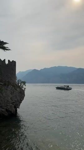 The video shows a serene lake scene with a ferry boat moving across the water. On the left side of the frame, there's a rocky cliff with a stone structure that appears to be part of a historical building, possibly a tower or fortification. A lone tree grows from the cliff face. The lake stretches out to the horizon, where a range of hazy blue mountains can be seen. The sky is overcast, with a hint of sunlight breaking through at the top right of the frame. The water is calm, with gentle ripples. The ferry boat is dark blue and white with windows, and there appear to be people on board. The overall atmosphere is peaceful and picturesque.