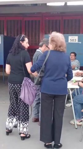 The scene shows a group of people gathered around a table outside a building, seemingly engaged in some sort of administrative or electoral process. There are four people standing near the table. The woman nearest the camera is wearing a black shirt and black pants with a white floral pattern. She has a purple shoulder bag. Next to her is a man in glasses, wearing a gray sweater and pants. Beside him, there is a woman with short auburn hair wearing a navy blue top and black pants. She has a black shoulder bag. These three people appear to be waiting in line. A fourth person is partially visible in the background. Five people sit around the table. One is a man wearing a striped shirt. Another four people sit at the table and seem to be assisting. There is a sign on the table reading “MESA 503.” The building behind has red gates and white walls. A sign can be seen above the gate. Further up, a red sign is also visible with the word “TOR” and "DES." The setting appears to be outdoors, possibly at a polling station or a registration center.