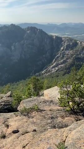 The video is a vertical shot of a mountainous landscape. In the foreground, there are large granite rocks with sparse vegetation. The middle ground features a steep, tree-covered slope descending into a valley. Further in the distance, rugged, rocky mountains rise under a clear blue sky. The scene is illuminated by natural sunlight, suggesting it is daytime. There are no people or man-made objects visible.